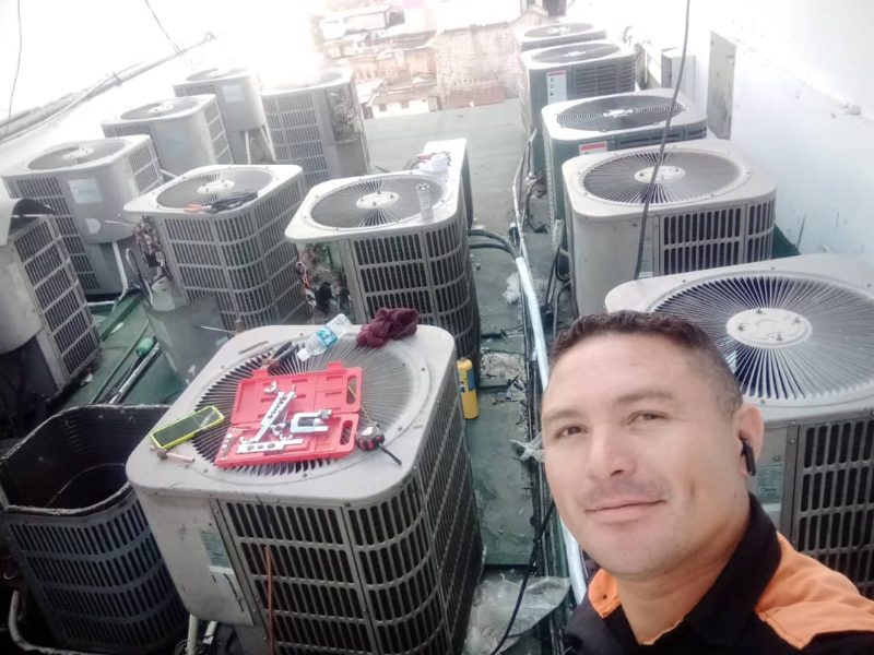 Un técnico en la azotea de un edificio, rodeado de una docena de condensadoras de aire acondicionado, con su maletín de herramientas de refrigeración sobre uno de los equipos.
